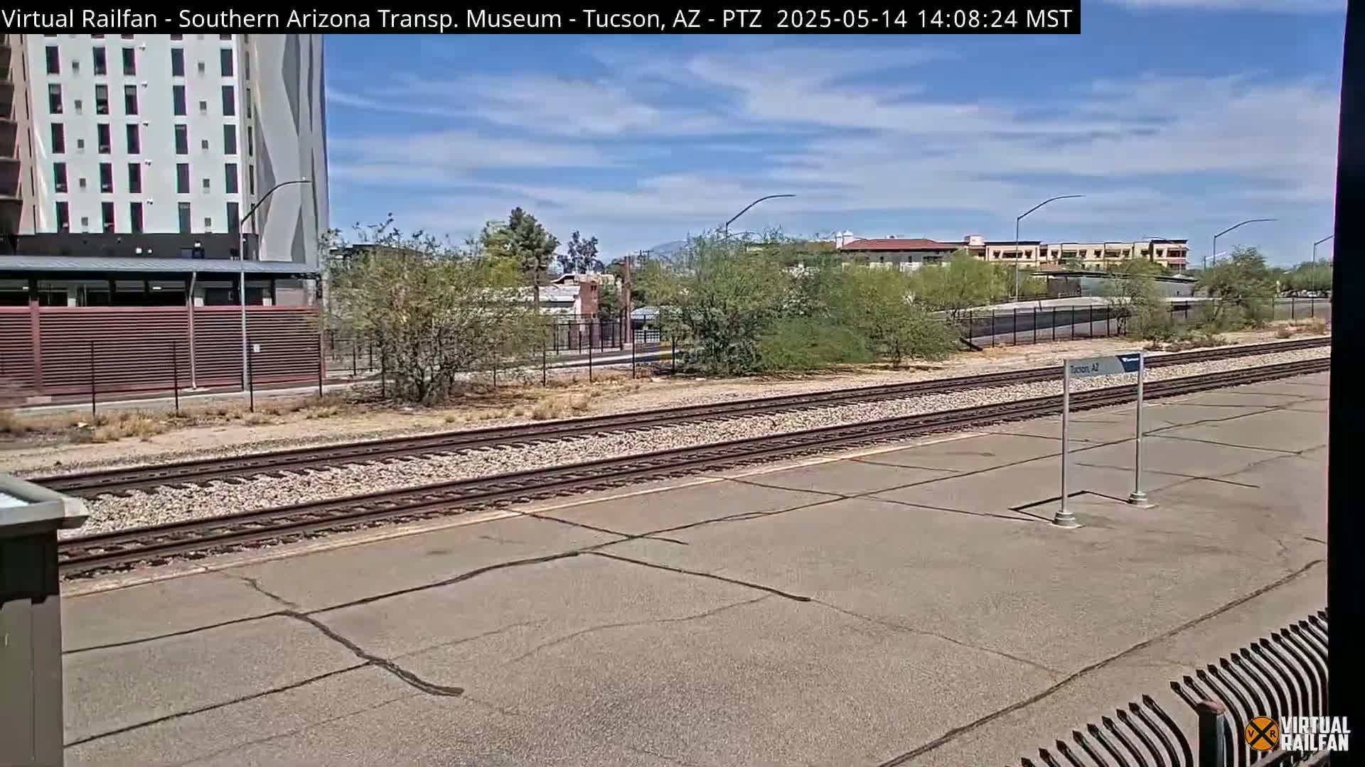 A mostly sunny day shows train tracks and a station platform next to a multi-story building and some shrubs.