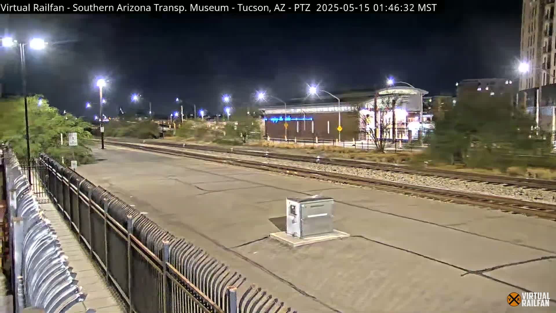 At night, a train track runs alongside a paved area next to a fenced-in platform with streetlights illuminating the scene.