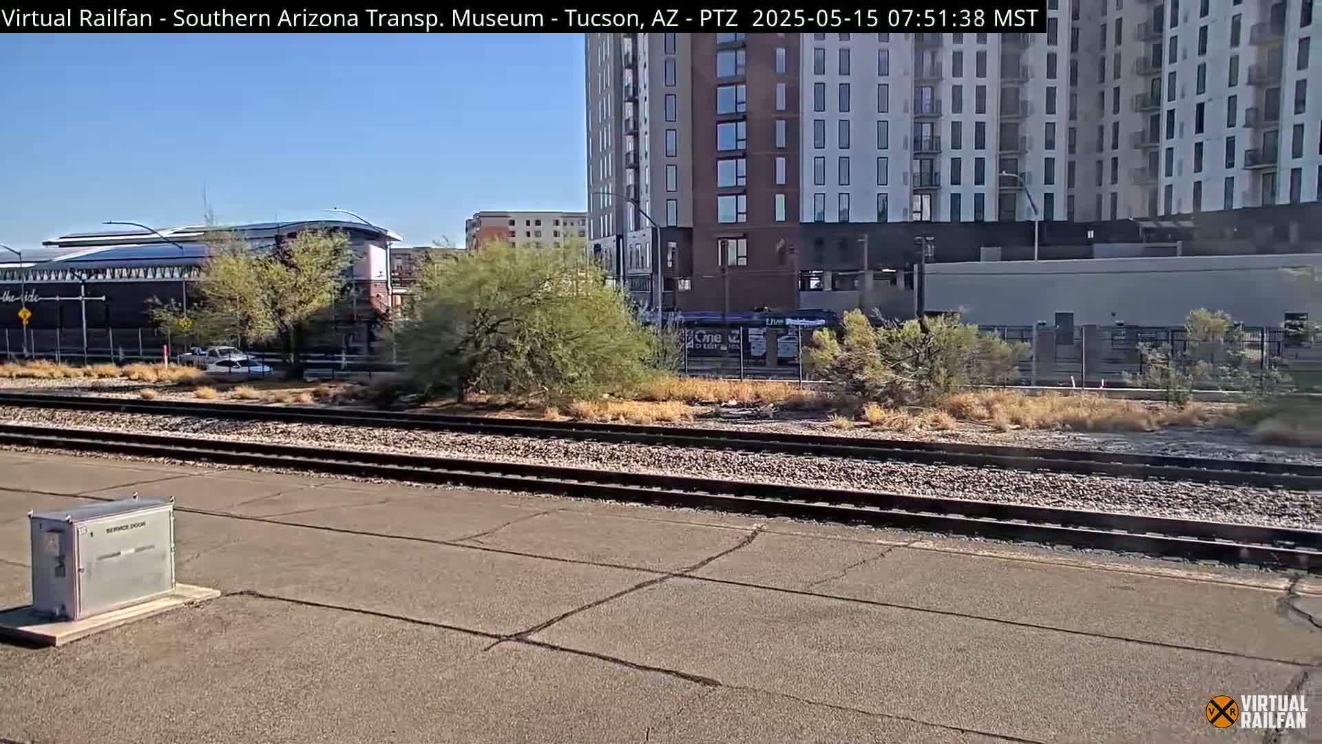 A sunny view of train tracks, a modern building, and desert vegetation.