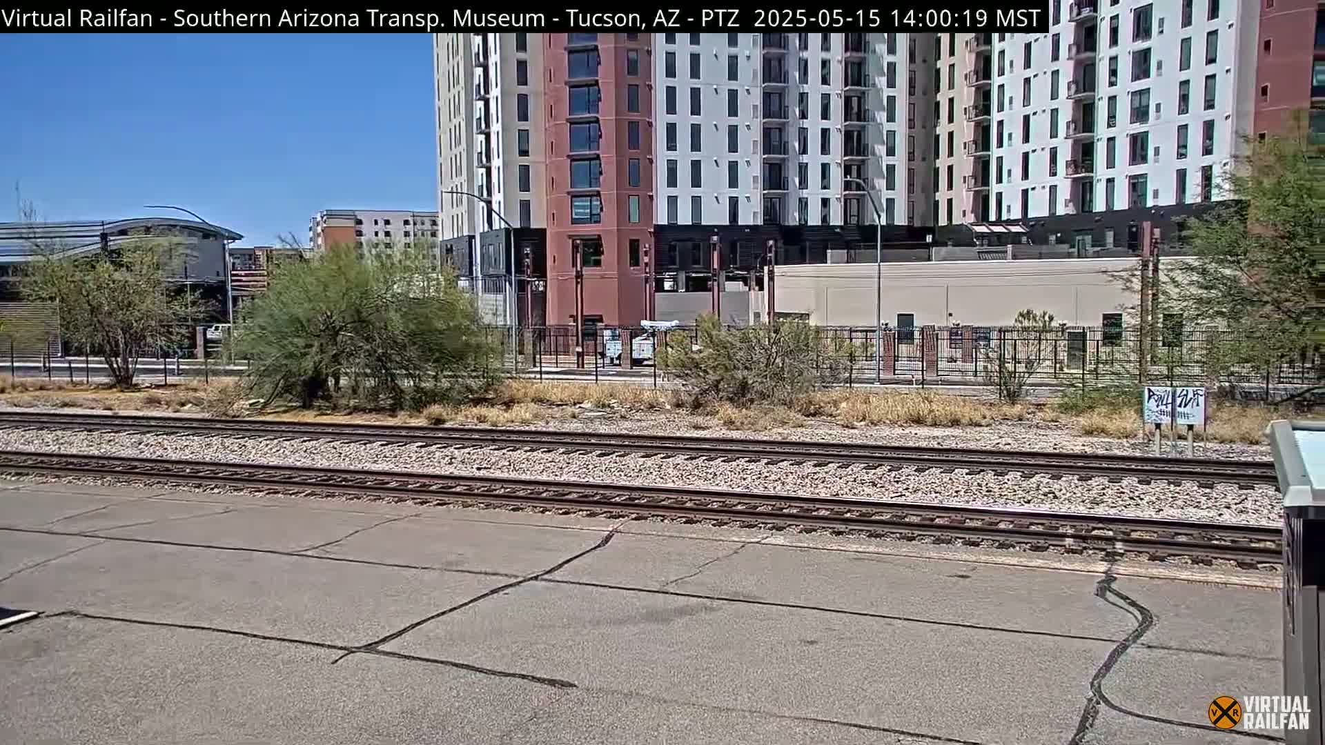 A sunny day view of railroad tracks in front of a multi-story building and sparse desert vegetation.