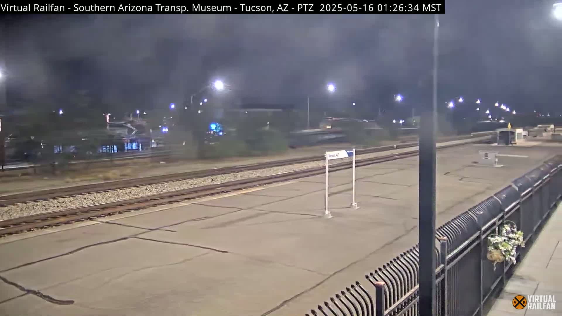 A nighttime view of a train station platform with train tracks, a few lights, and a metal fence with a hanging planter.