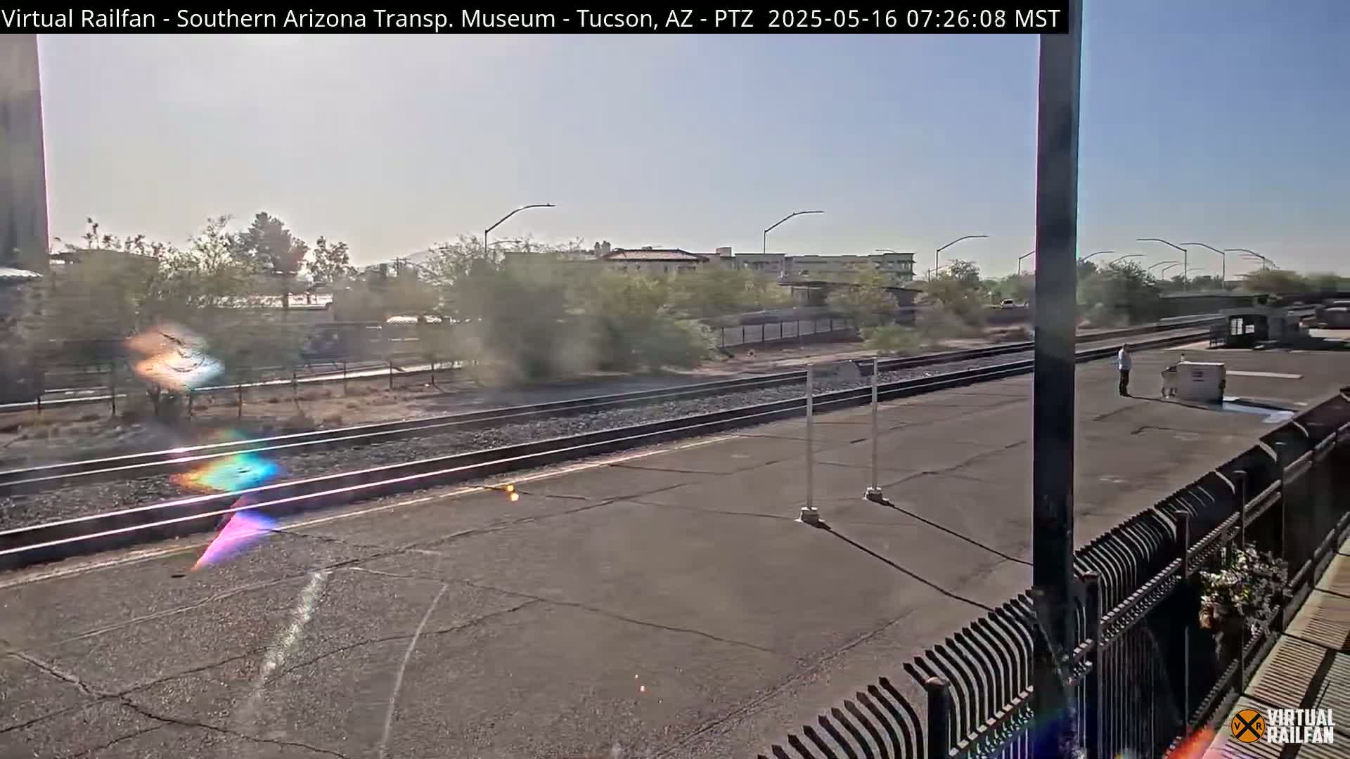 A sunny, hazy day at a train station with a person standing on the platform near a rail line and several buildings in the background.