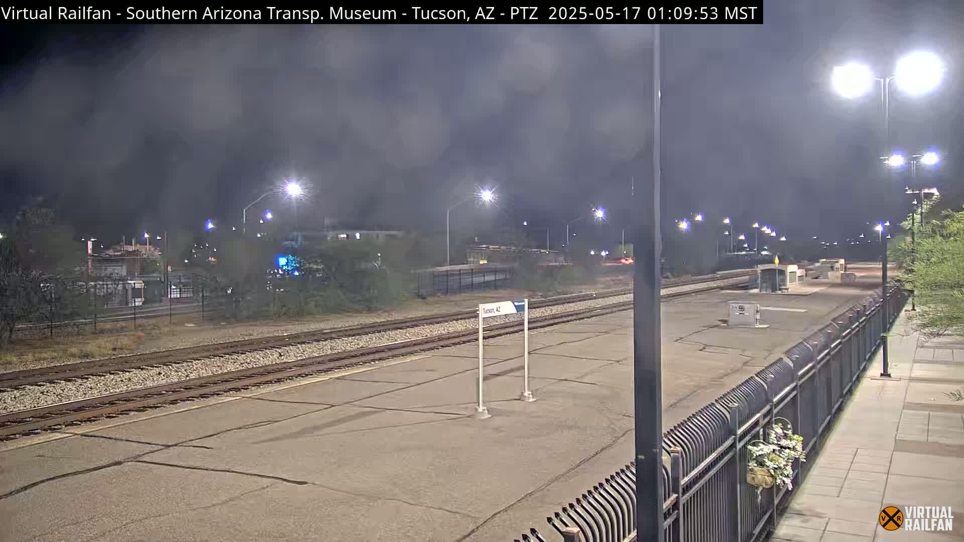 A nighttime view of a train station platform with train tracks, lights, and a fence under a hazy sky.