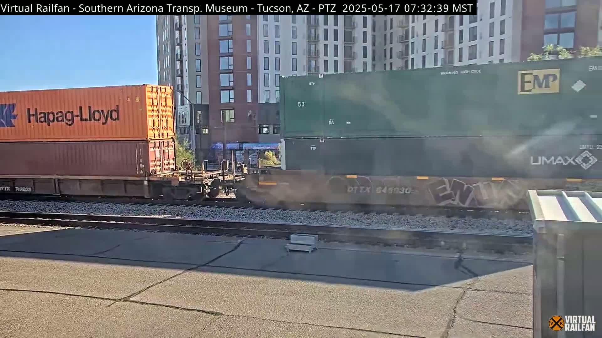 A freight train carrying shipping containers passes by on a sunny day next to a modern apartment building.