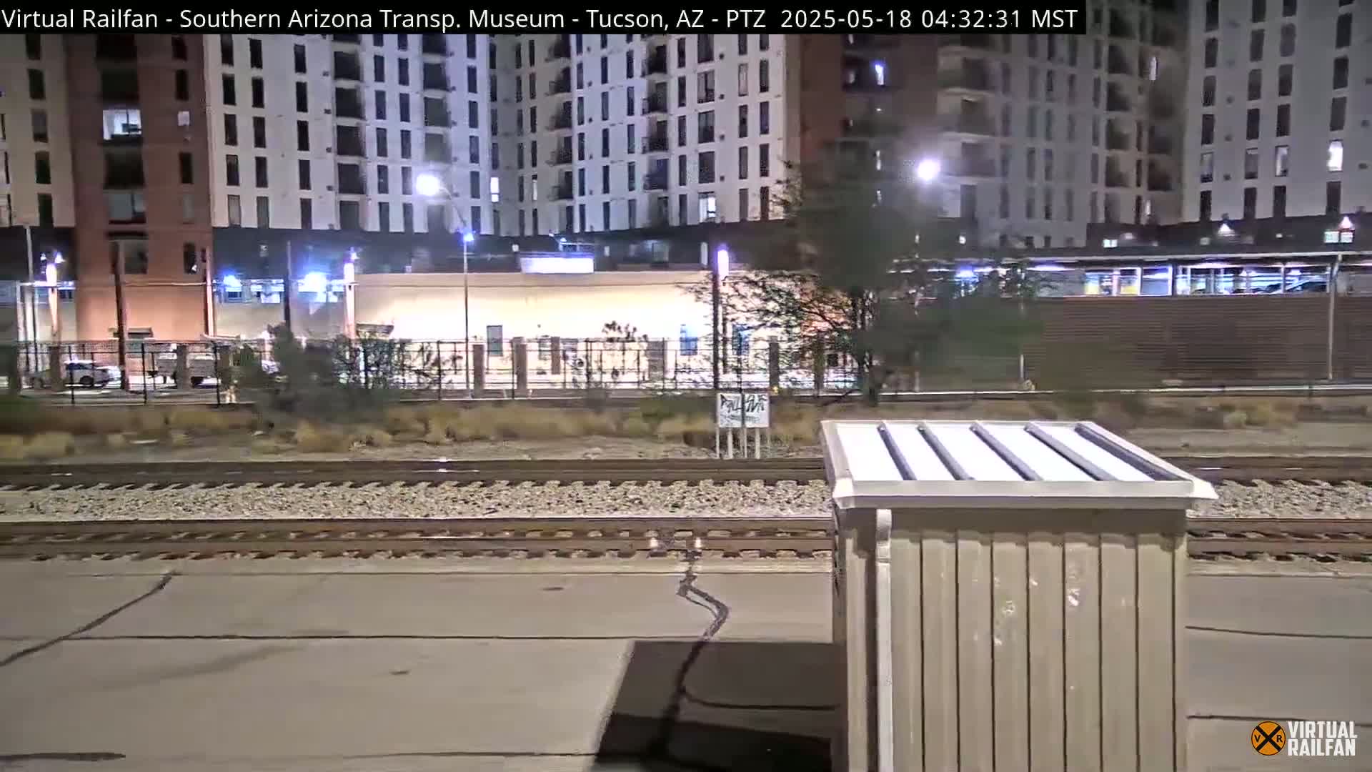 At night, a view of train tracks runs in front of a multi-story building under artificial light.