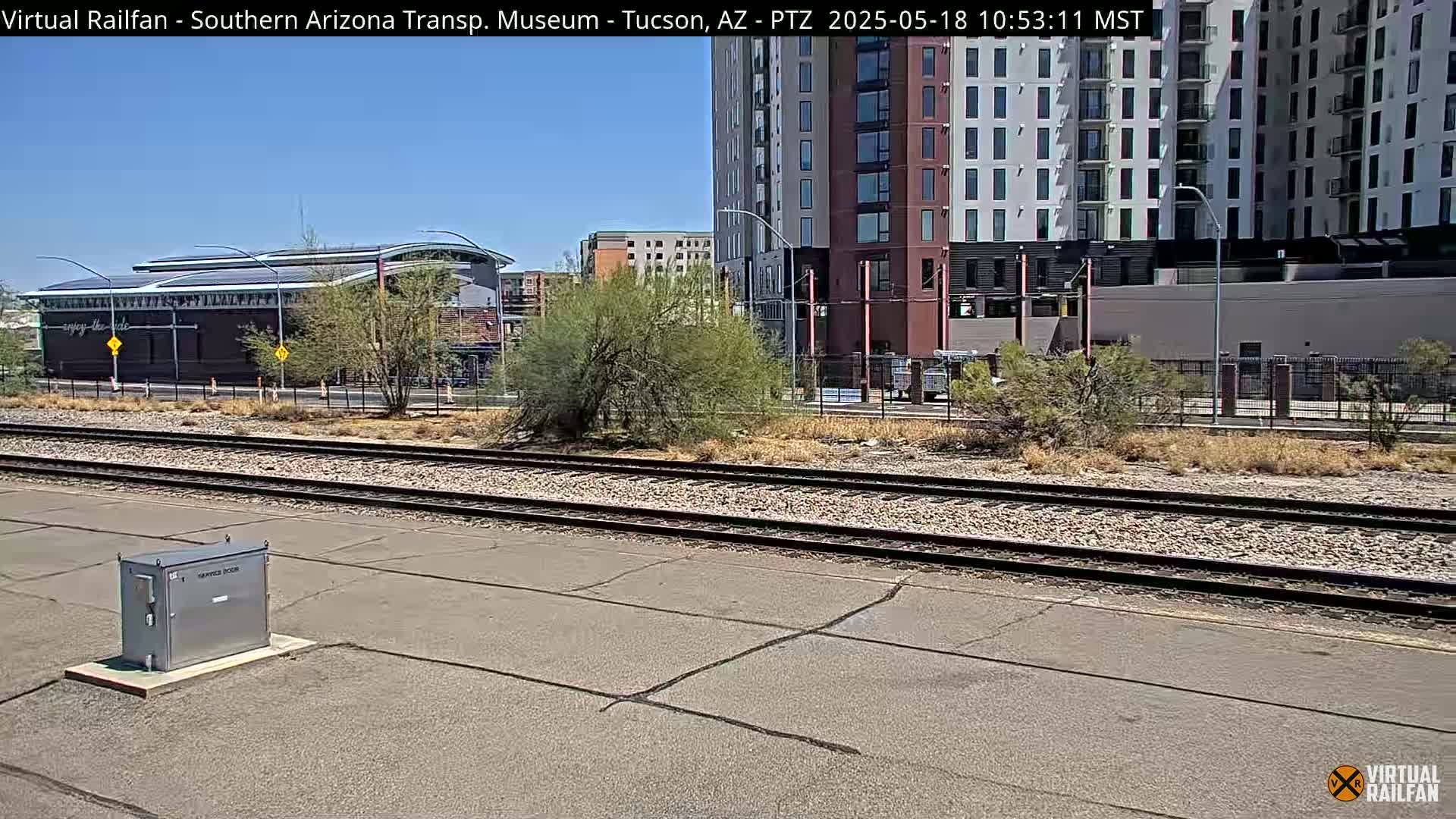 A sunny day shows a view of railroad tracks, a metal box, desert vegetation, and a multi-story building.