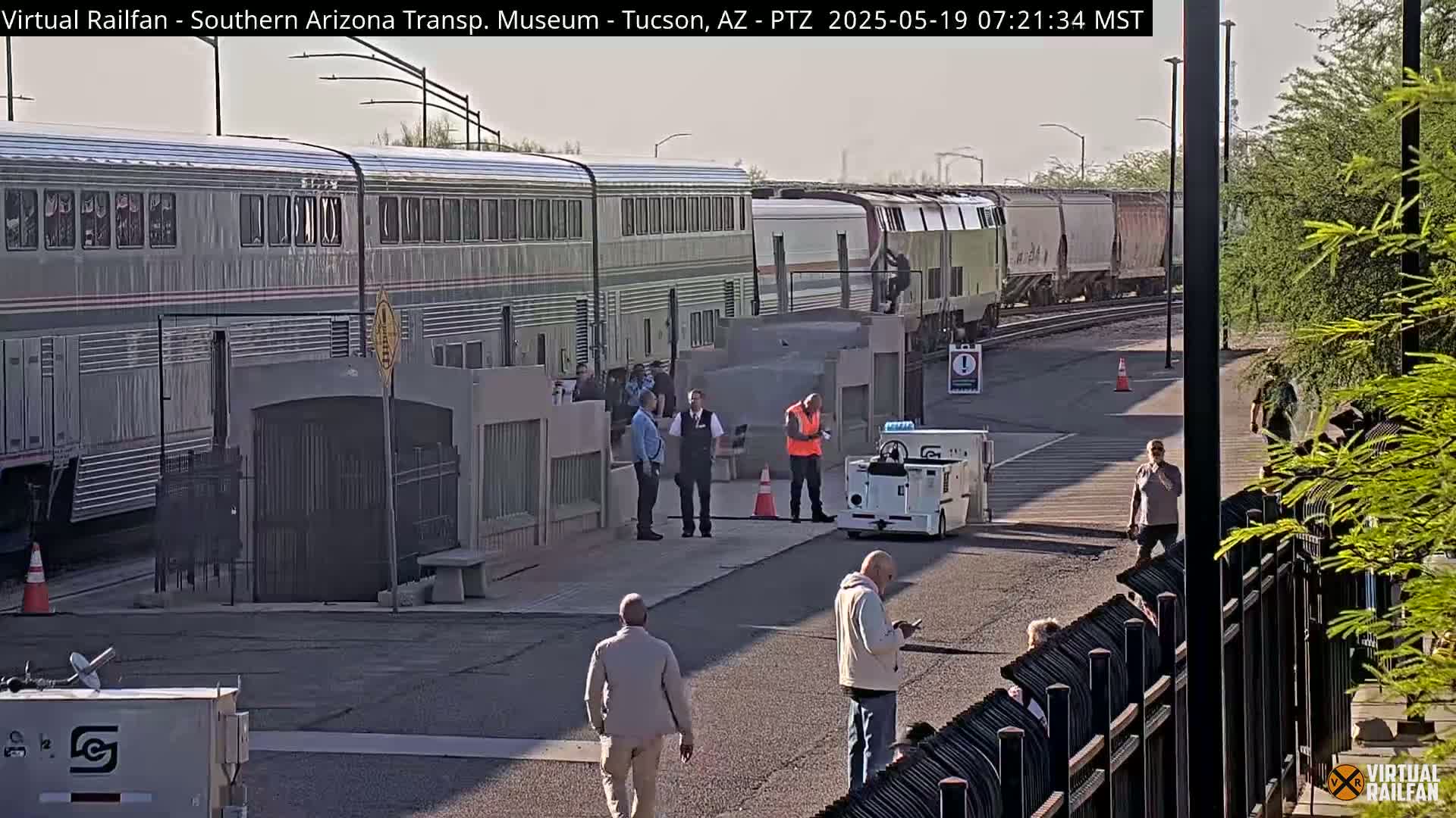 A long passenger train is parked at a station on a sunny day, with several people standing nearby and a baggage cart in the foreground.