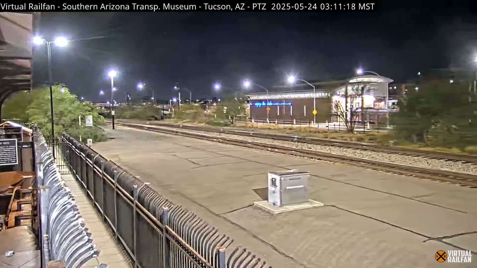 A nighttime view of a train station platform with train tracks extending into the distance under bright artificial lights.