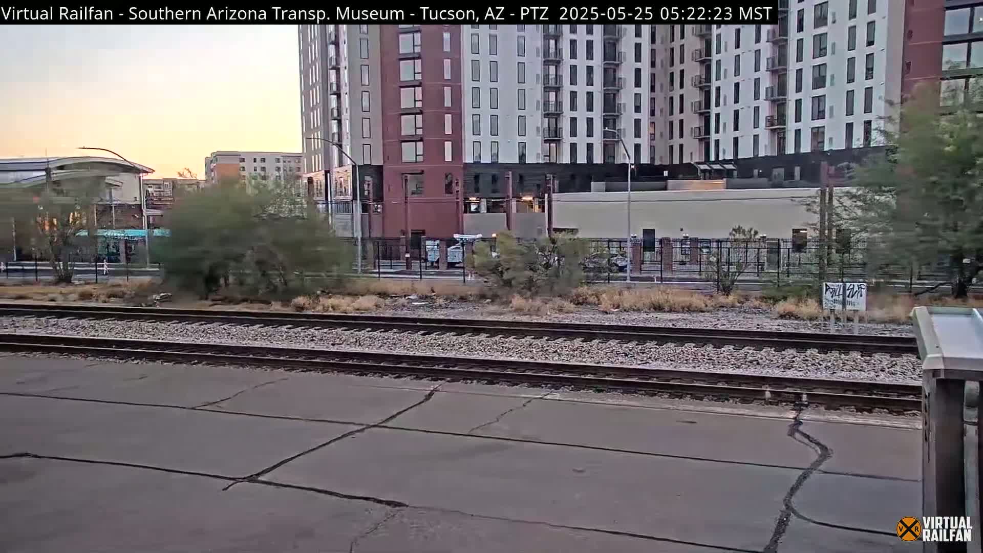 A view of railroad tracks next to a paved area, with a tall apartment building and desert vegetation in the background under a clear, dawn sky.