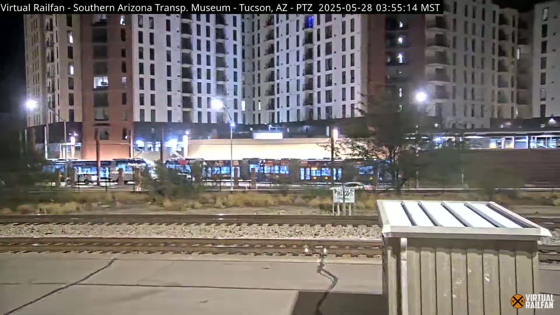 At night, several light rail vehicles are parked at a station in front of a tall apartment building.