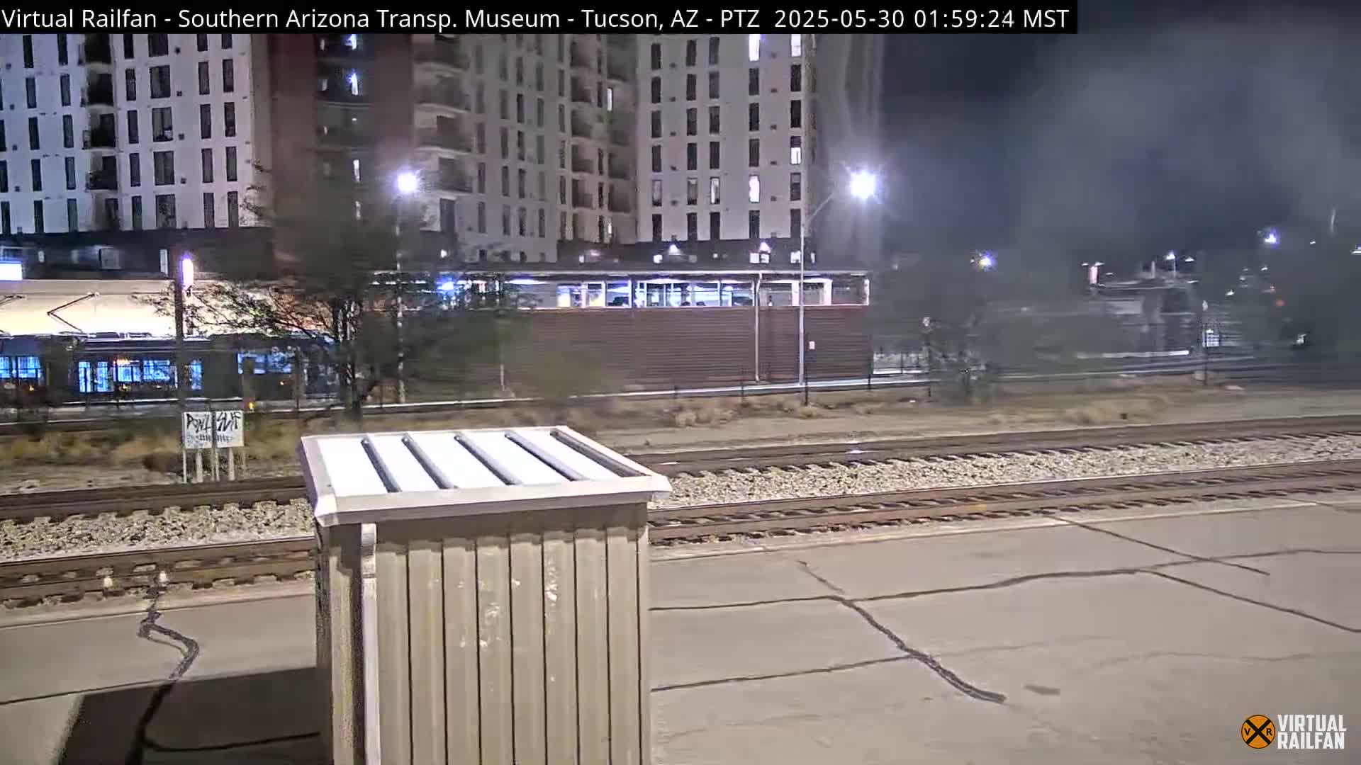 A nighttime view of a train station with a light-brown utility box in the foreground, train tracks, and apartment buildings in the background under dark, clear skies.