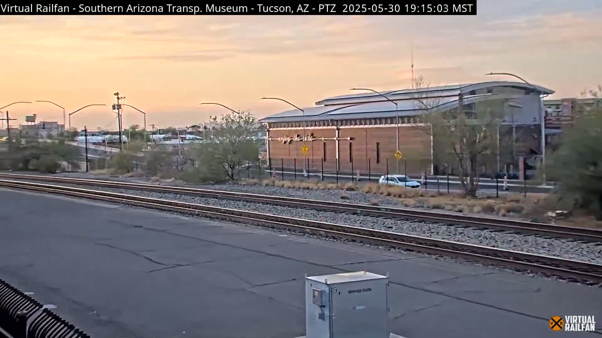 Under a twilight sky, a long, low building with a curved roof sits beside railroad tracks.