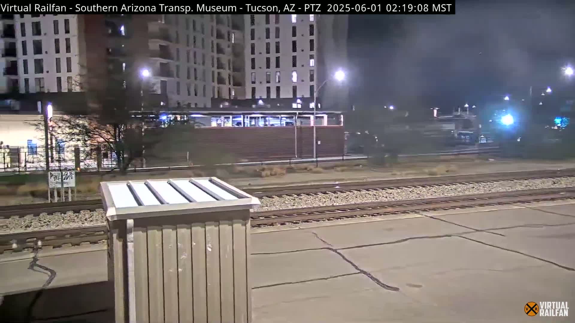 A nighttime view of train tracks at a station near a multi-story building, with a train in the distance under cloudy skies.