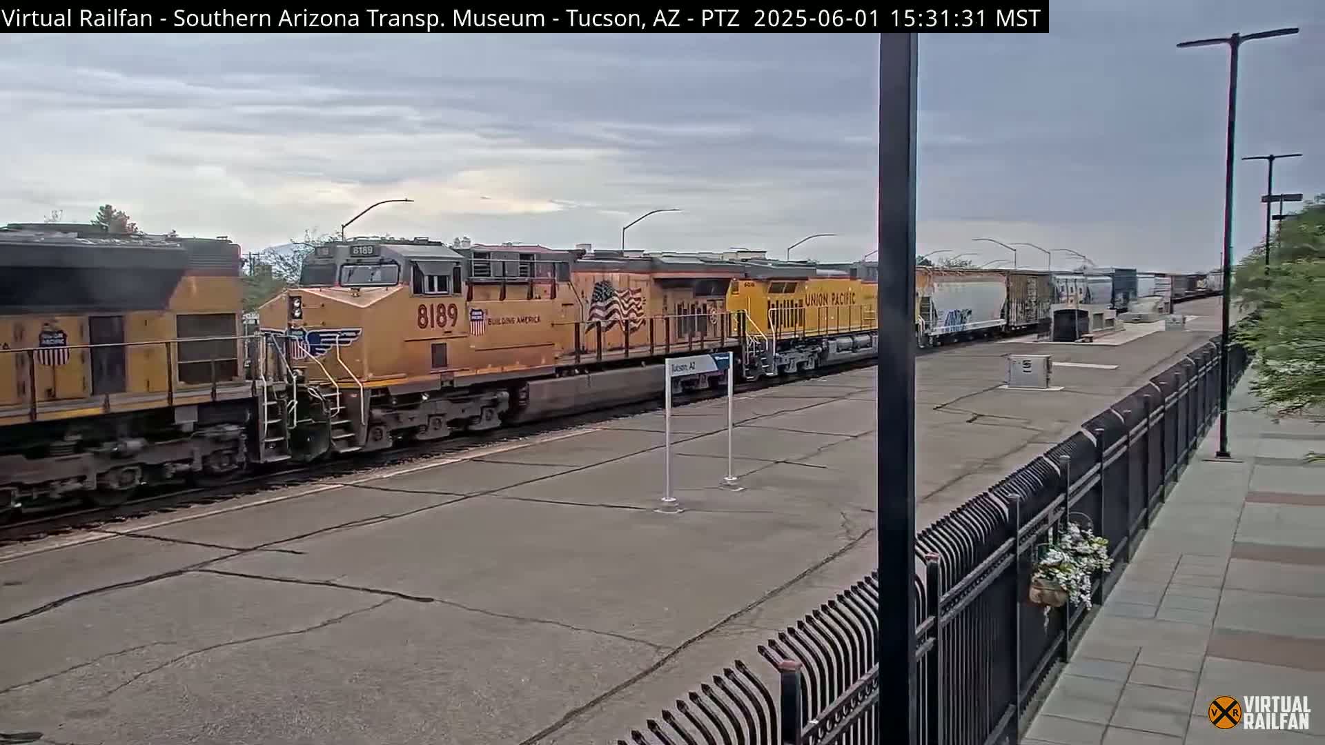 A long freight train of yellow locomotives and various cargo cars sits on tracks under an overcast sky next to a black metal fence.