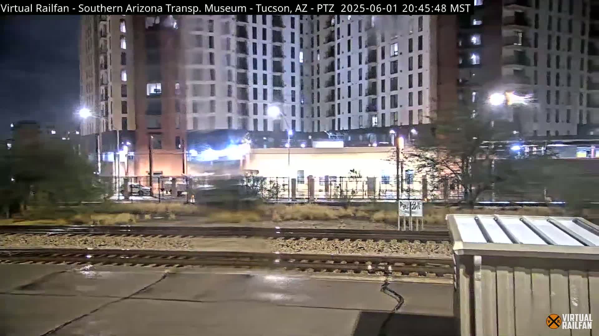 A train passes by a modern apartment building at night, under slightly wet conditions.