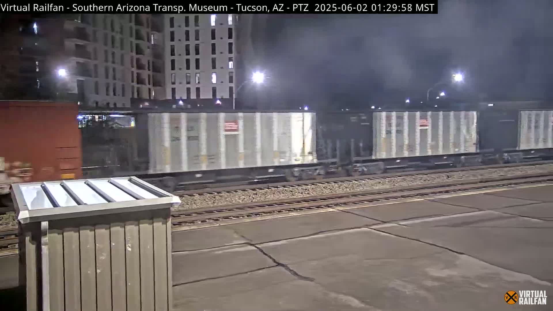 At night, a freight train passes by a rail yard next to a multi-story building.