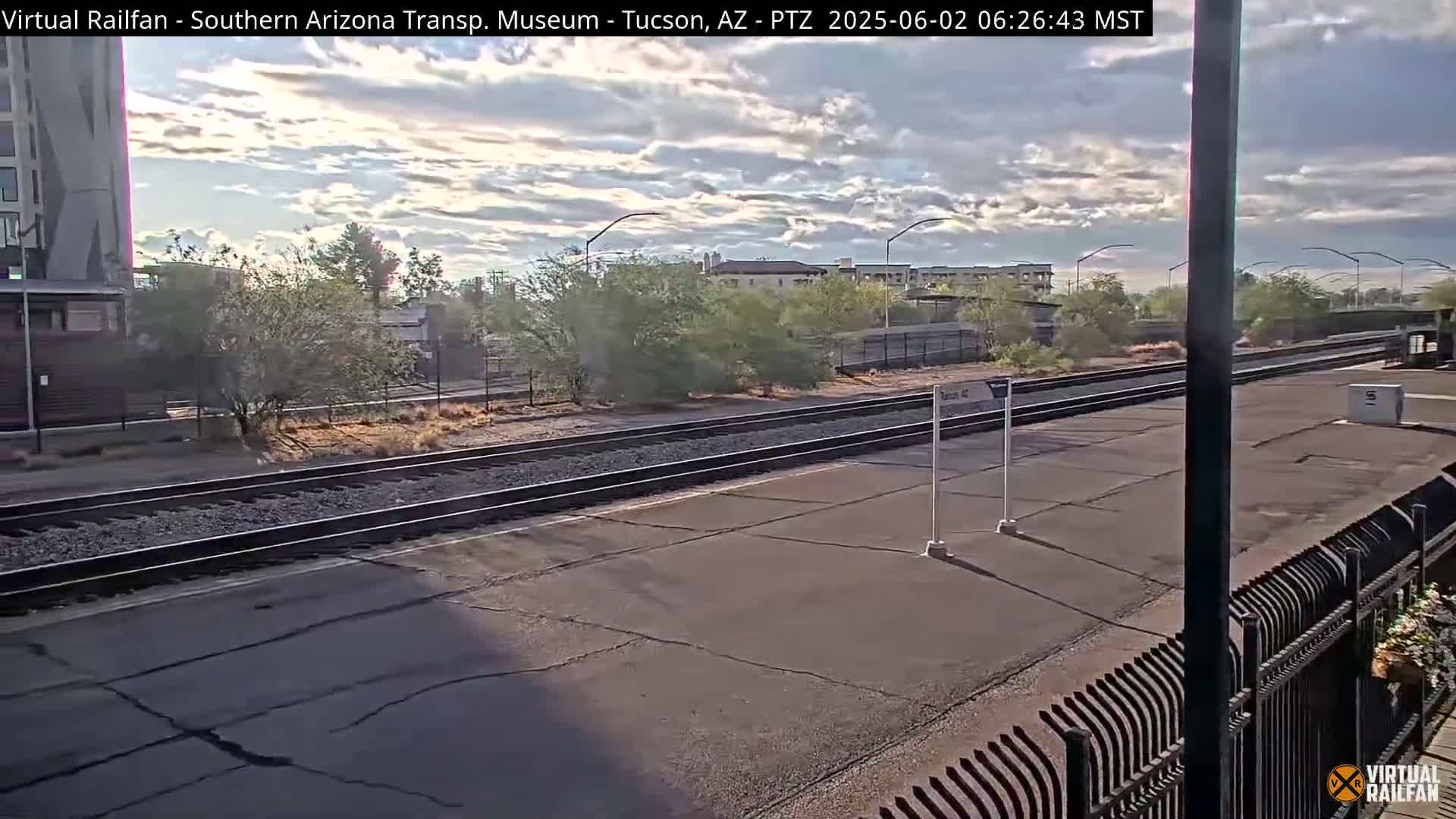A partly cloudy morning scene shows train tracks at a station, surrounded by desert landscaping and low buildings.