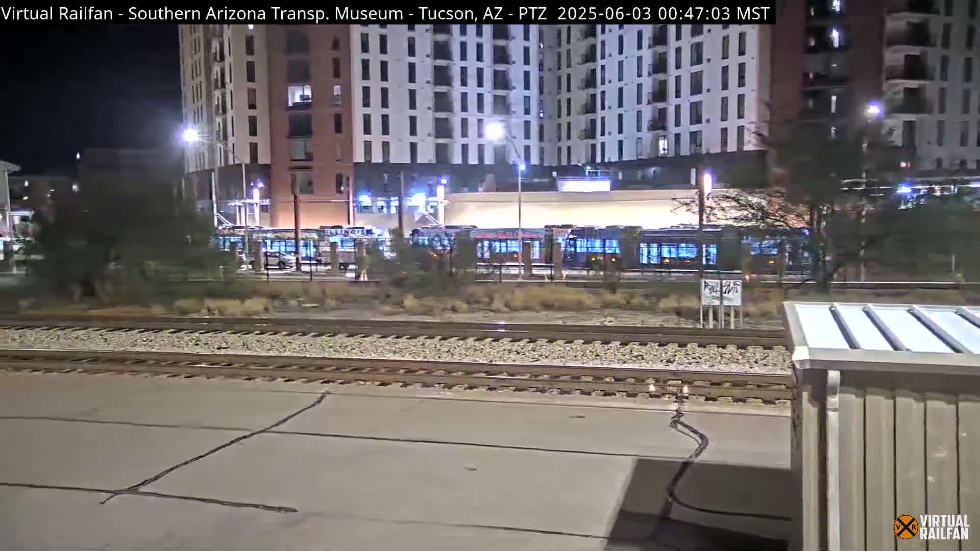 At night, several light-rail vehicles are parked near a multi-story building next to train tracks.