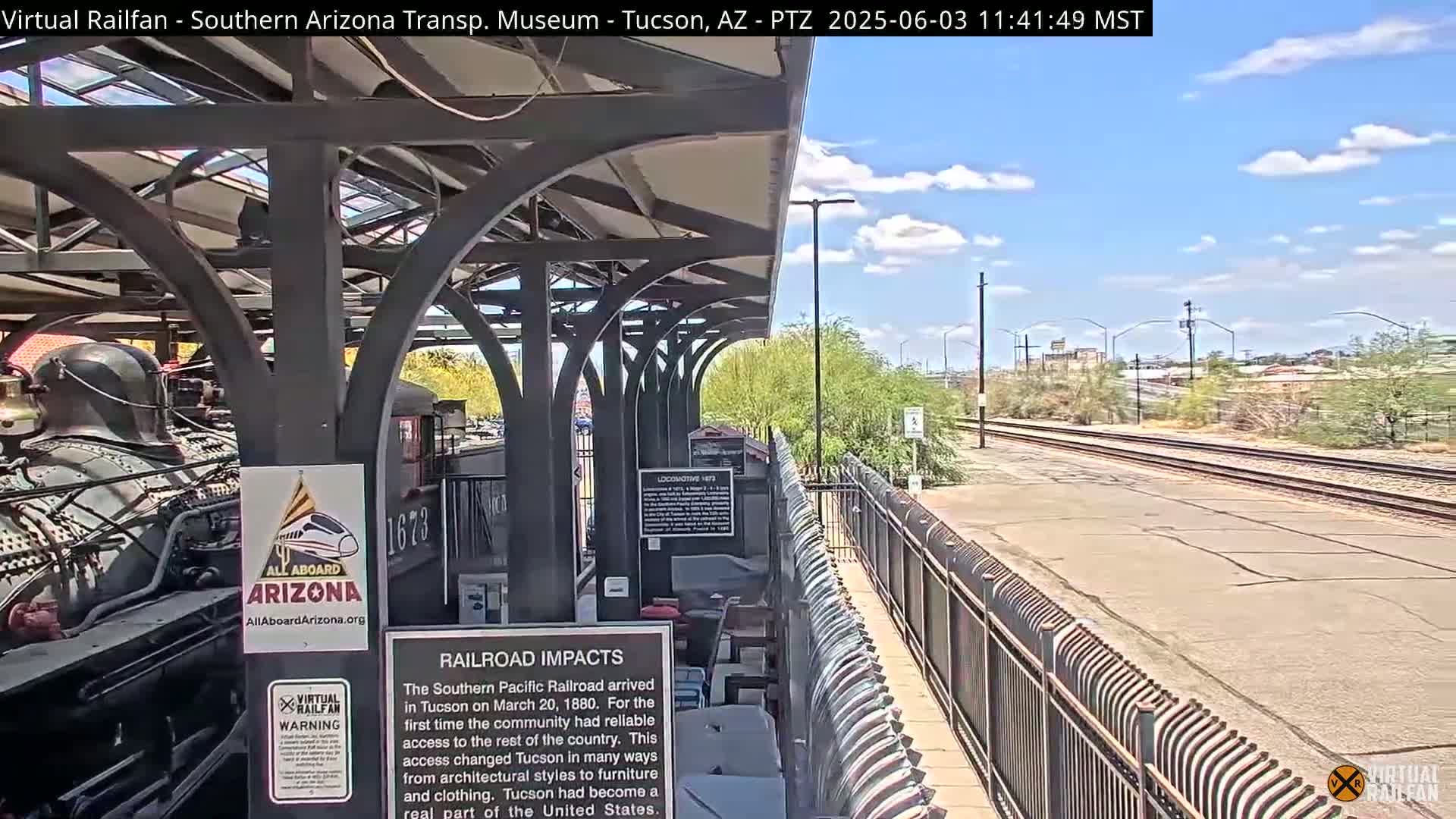 A partly cloudy day shows a train partially visible under a covered platform next to railroad tracks.