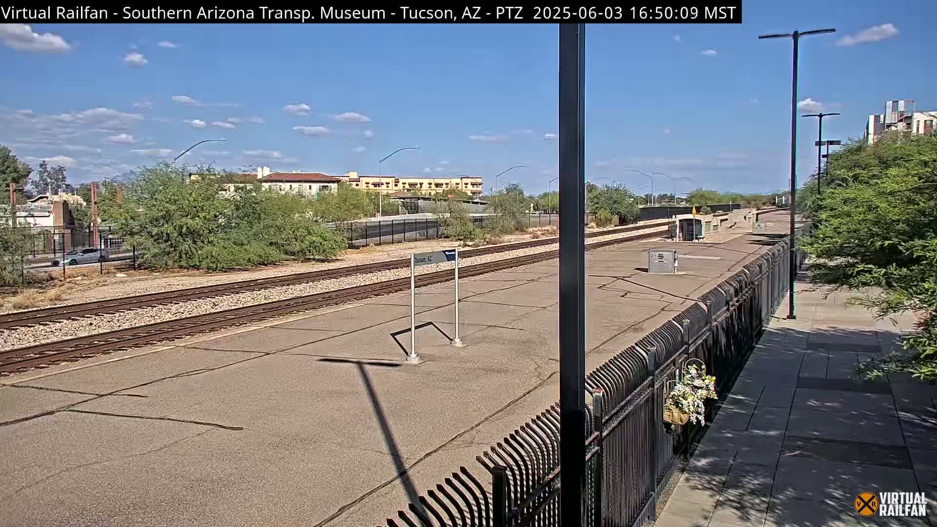 A sunny day at a train station with a paved platform, train tracks, a black metal fence, and some shrubs and buildings in the background.