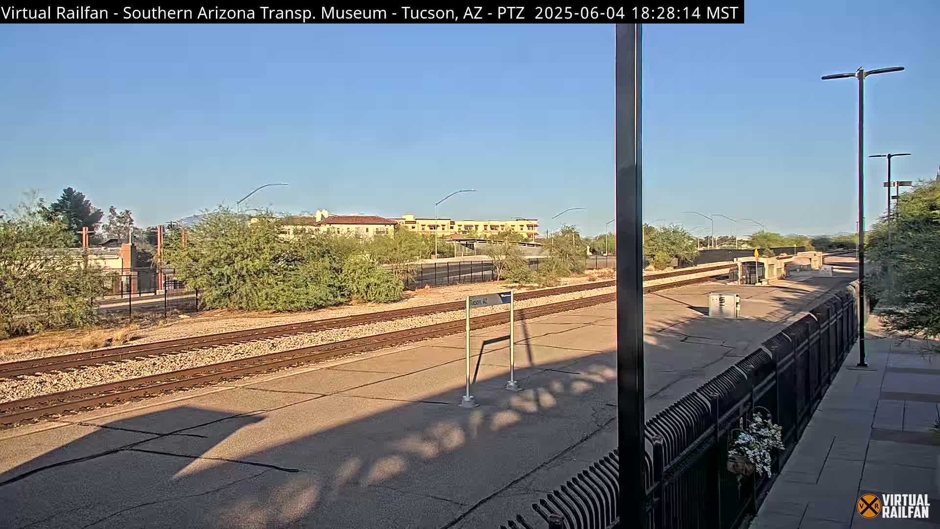 A sunny daytime view of train tracks and a station platform, with low desert vegetation and buildings in the background.