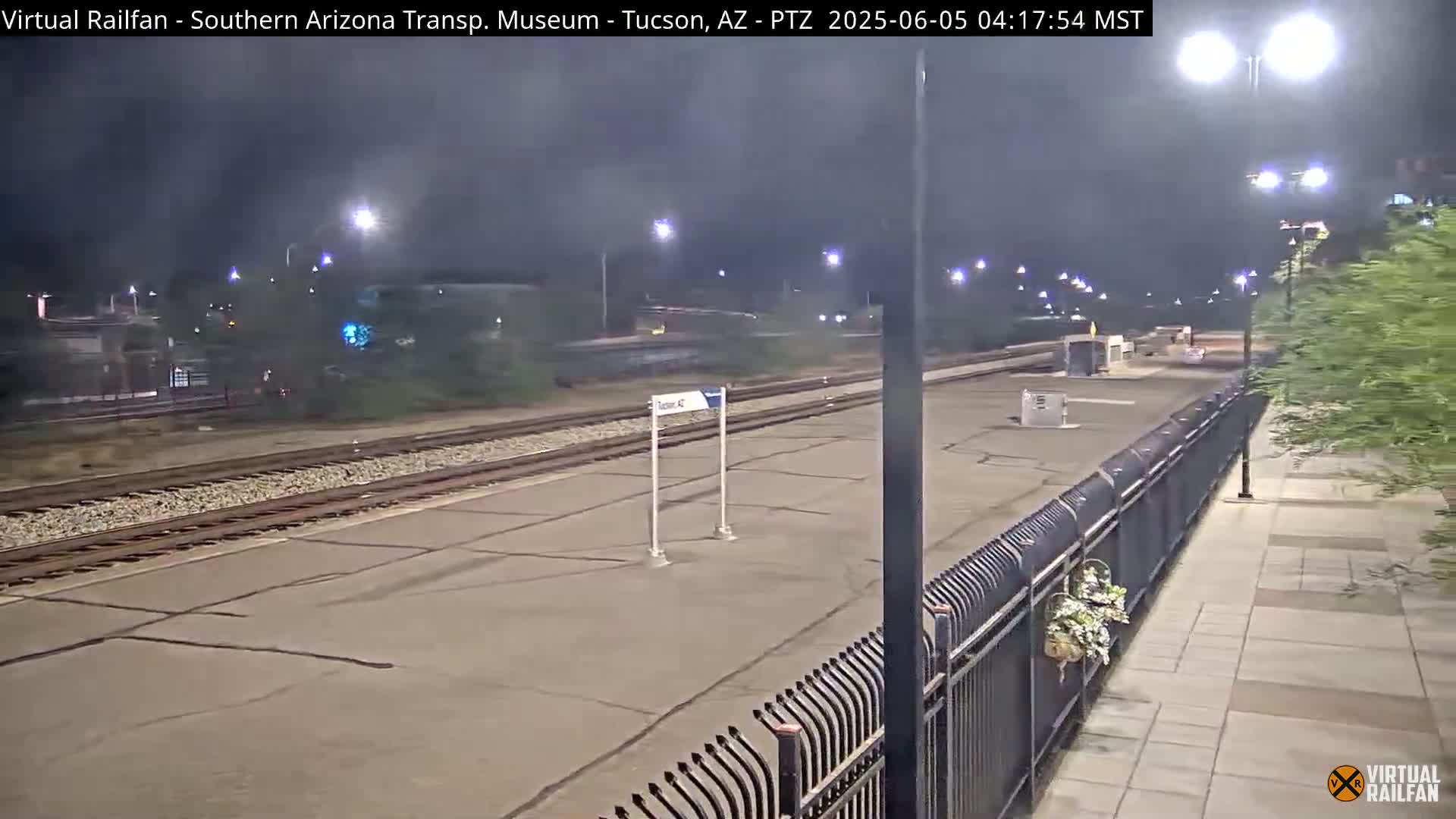 A nighttime view of a train station platform with railroad tracks, a fence, and some landscaping.