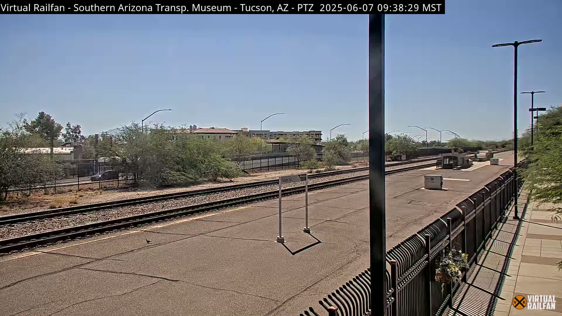 A sunny day at a train station with railway tracks extending into the distance, passing under a pedestrian overpass near a residential area.