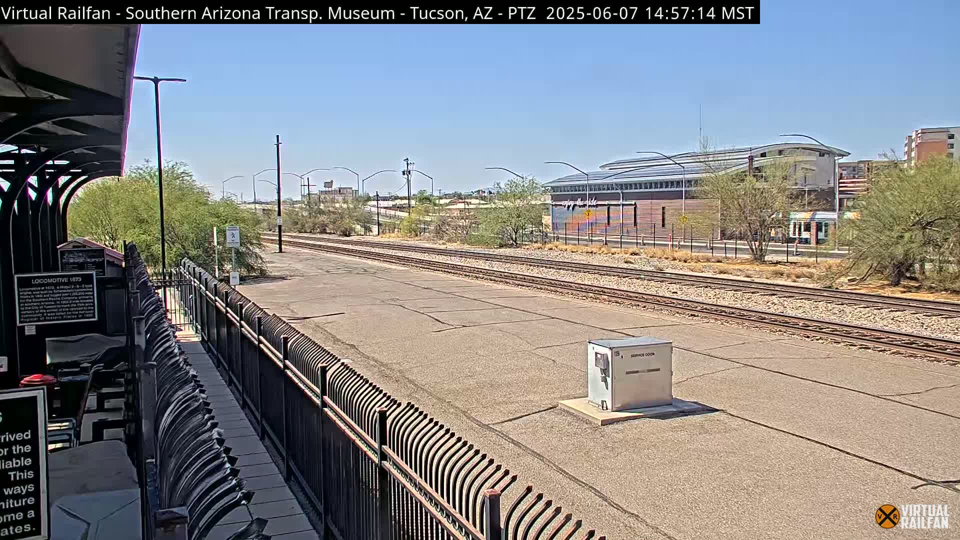 A sunny day view of a train station platform, railway tracks stretching into the distance, and a building with colorful artwork in the background.