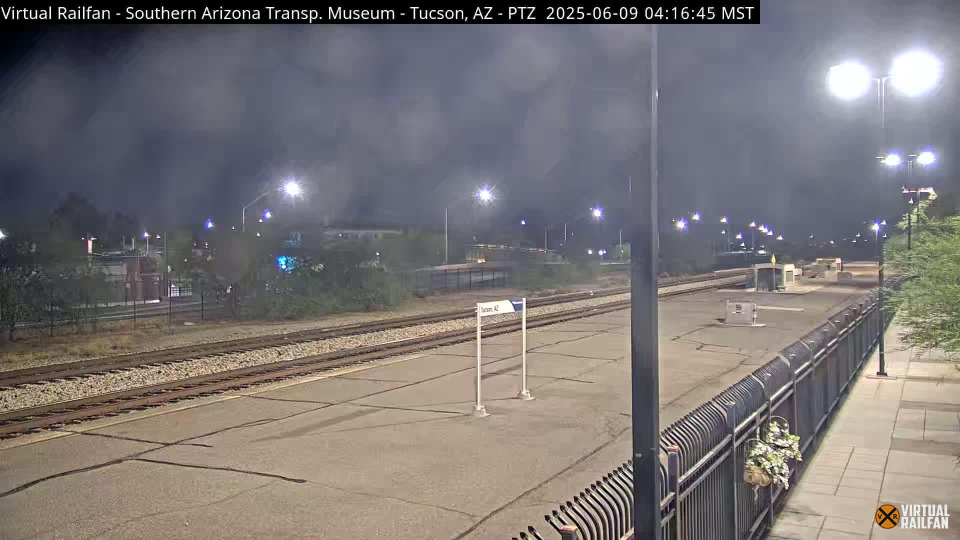 A nighttime view of a train station platform with multiple sets of train tracks, illuminated by streetlights under a cloudy sky.