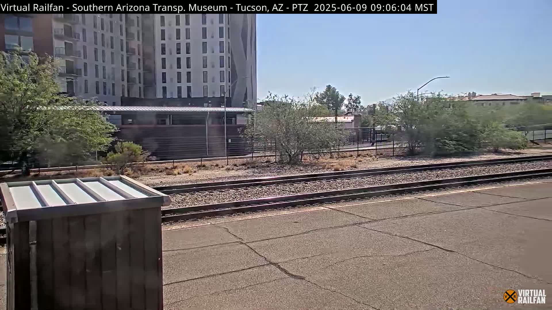 A view of train tracks on a sunny day, with a building and desert vegetation in the background.