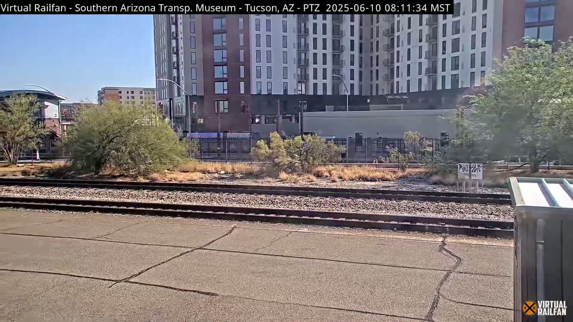 A view of train tracks next to a multi-story building and desert landscaping on a sunny day.