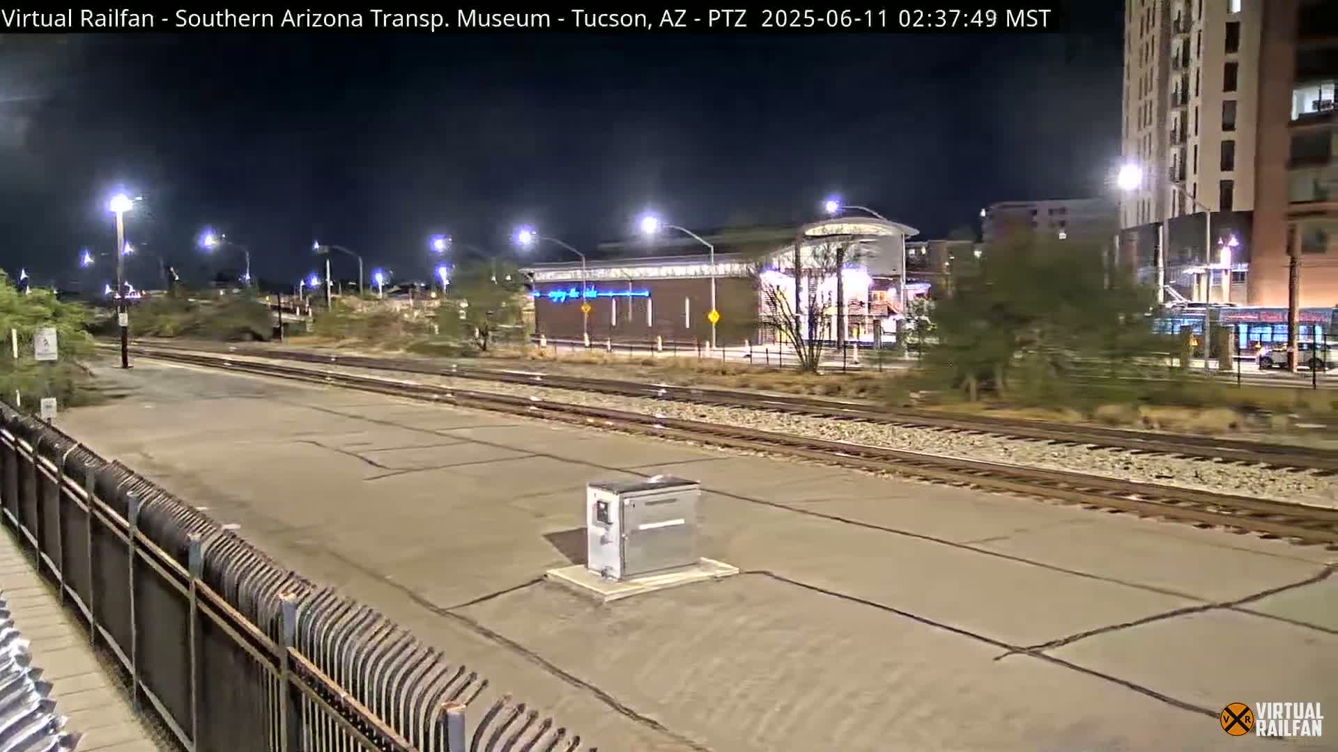 A nighttime view of a train station platform with train tracks, a metal fence, and a building in the background under clear skies.