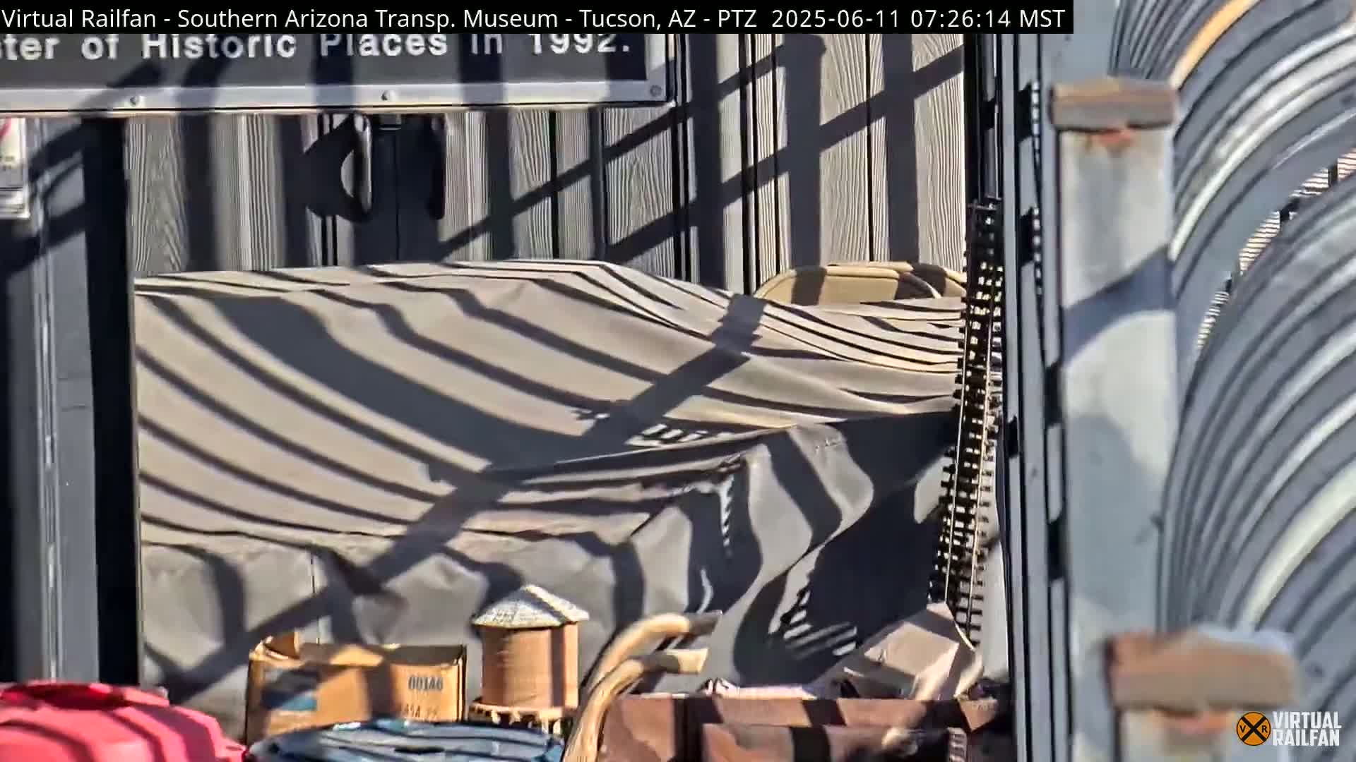 A tan tarp draped over something, possibly outdoor furniture, is partially visible in bright sunlight, casting striped shadows on the tarp.
