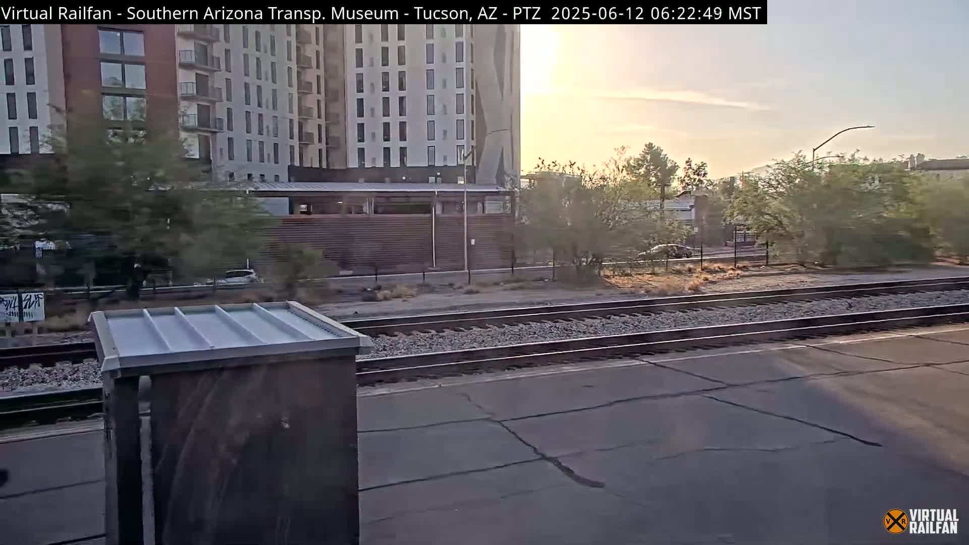 A sunrise view of a train track next to a multi-story building and sparse desert vegetation.