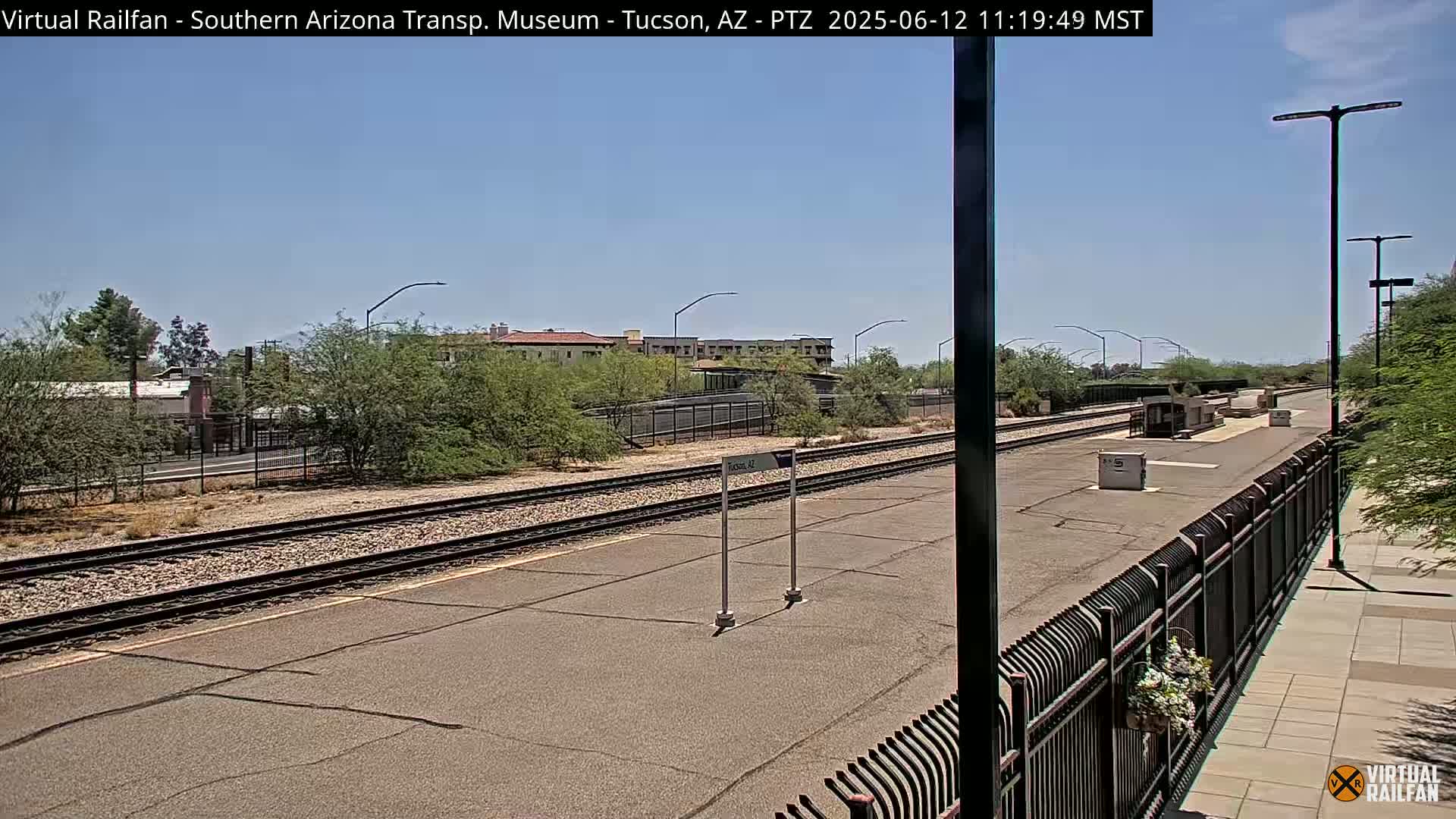 A train station platform with two sets of train tracks under a clear, sunny sky.