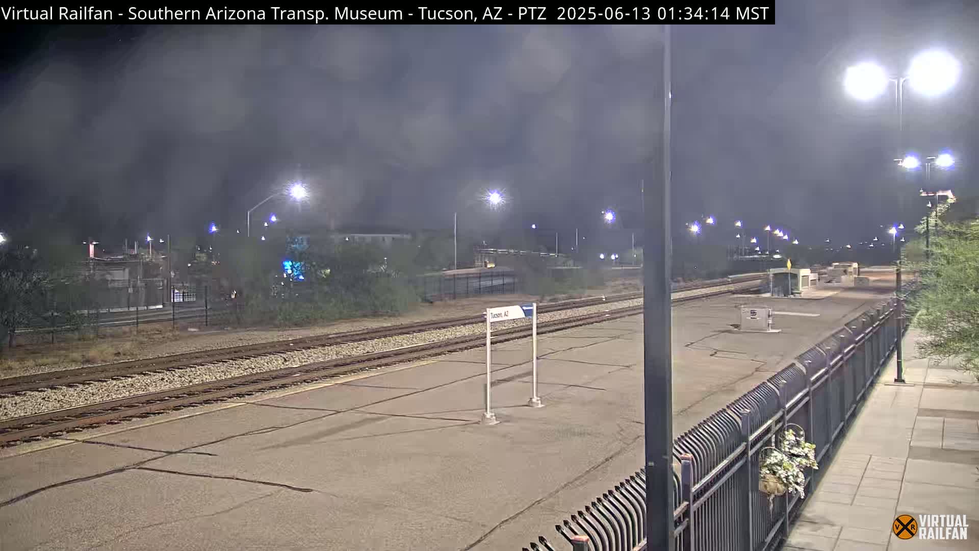 A nighttime view of a deserted train station platform with multiple tracks, illuminated by streetlights under a hazy sky.