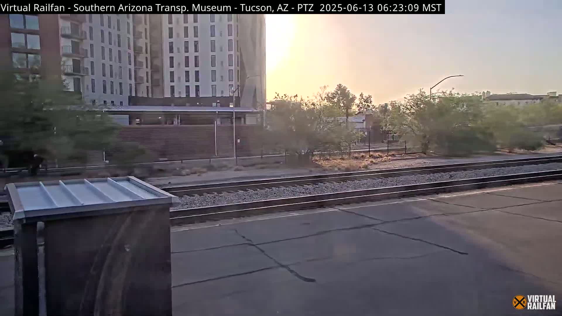 A sunrise view of train tracks next to a modern building and desert vegetation.