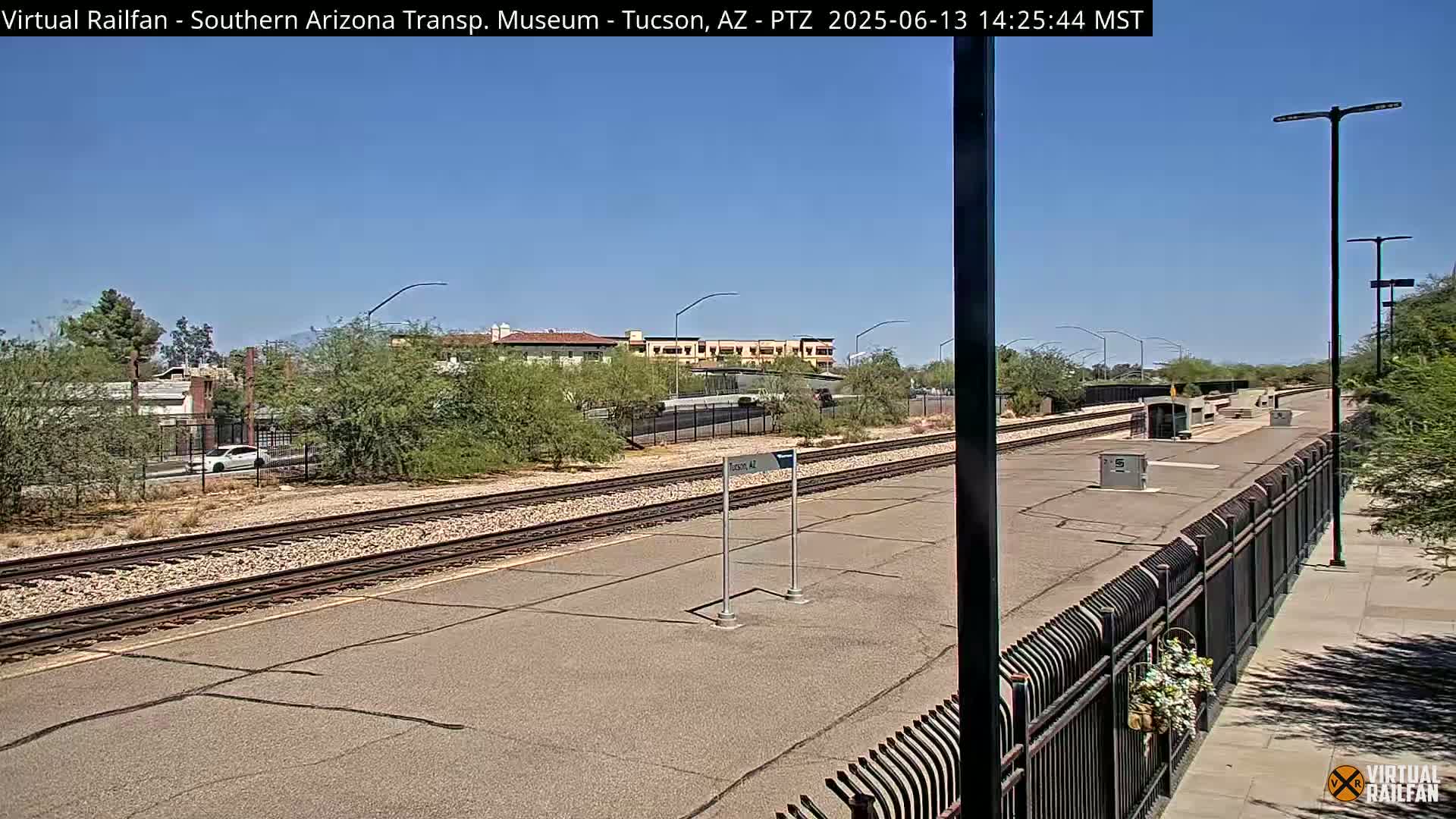 A sunny day at a train station with a paved platform, train tracks, and a metal fence, bordered by desert vegetation and buildings in the distance.