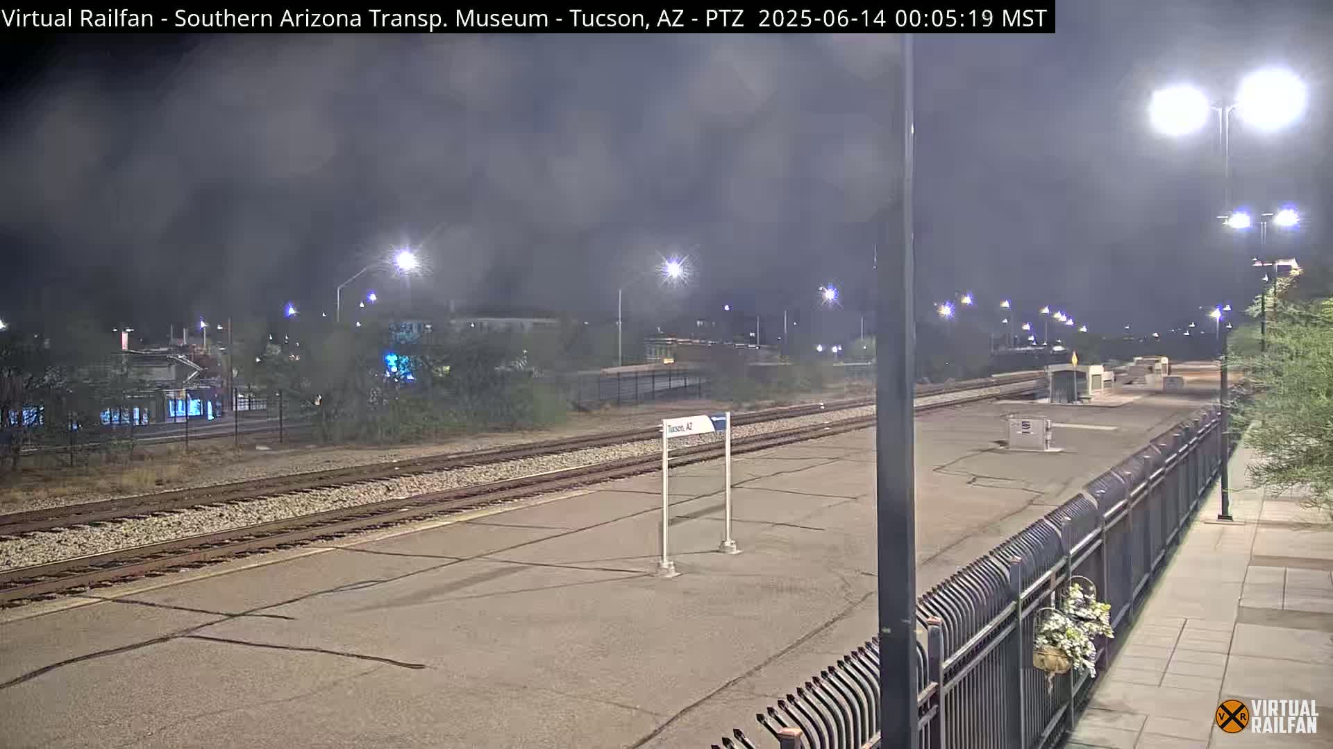 A nighttime view of a train station platform with train tracks, a fence, and streetlights under a cloudy sky.