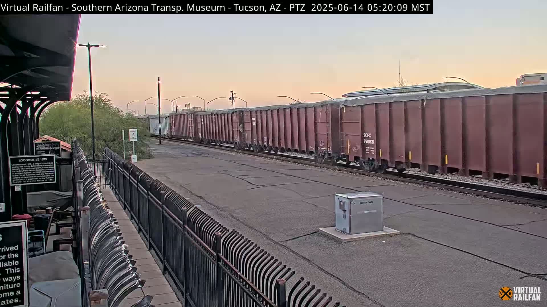 A long freight train of reddish-brown covered hopper cars passes by a train station under a pale dawn sky.