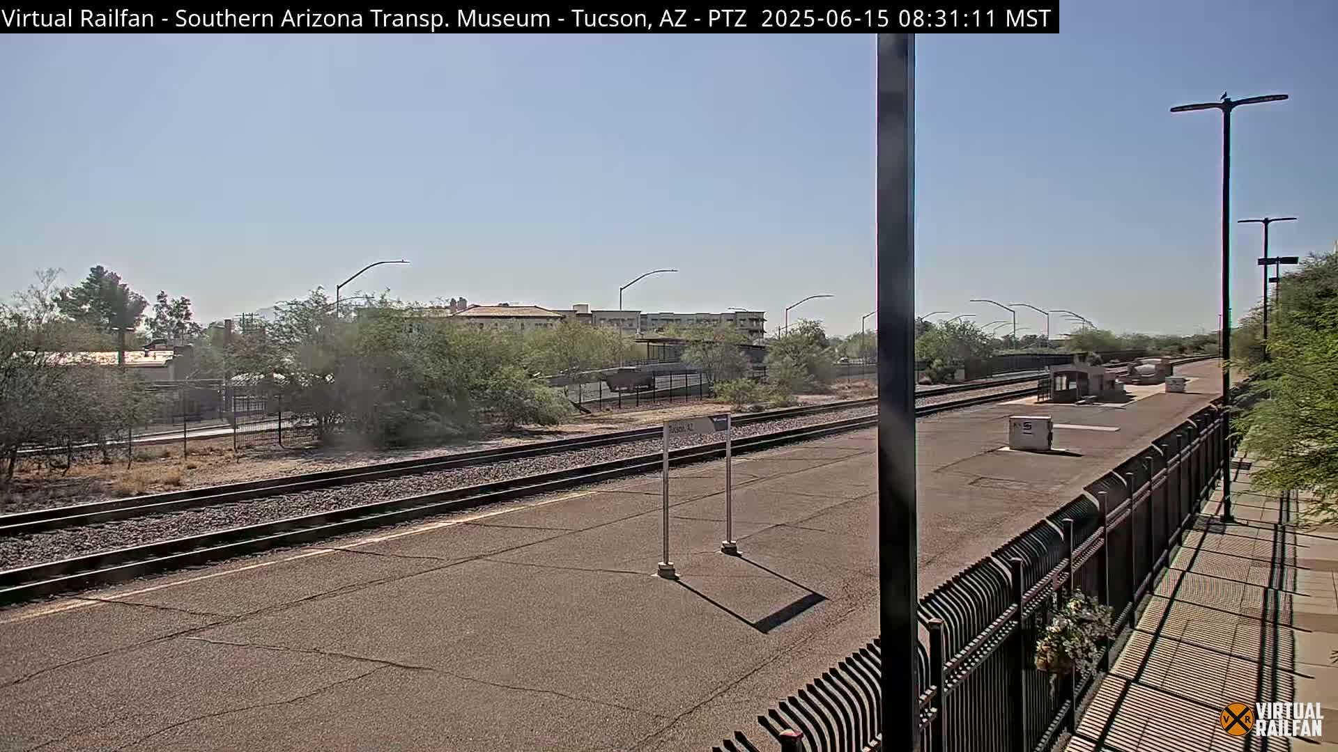 A sunny day at a train station with a paved platform, train tracks, and a chain-link fence separating the platform from a sidewalk.