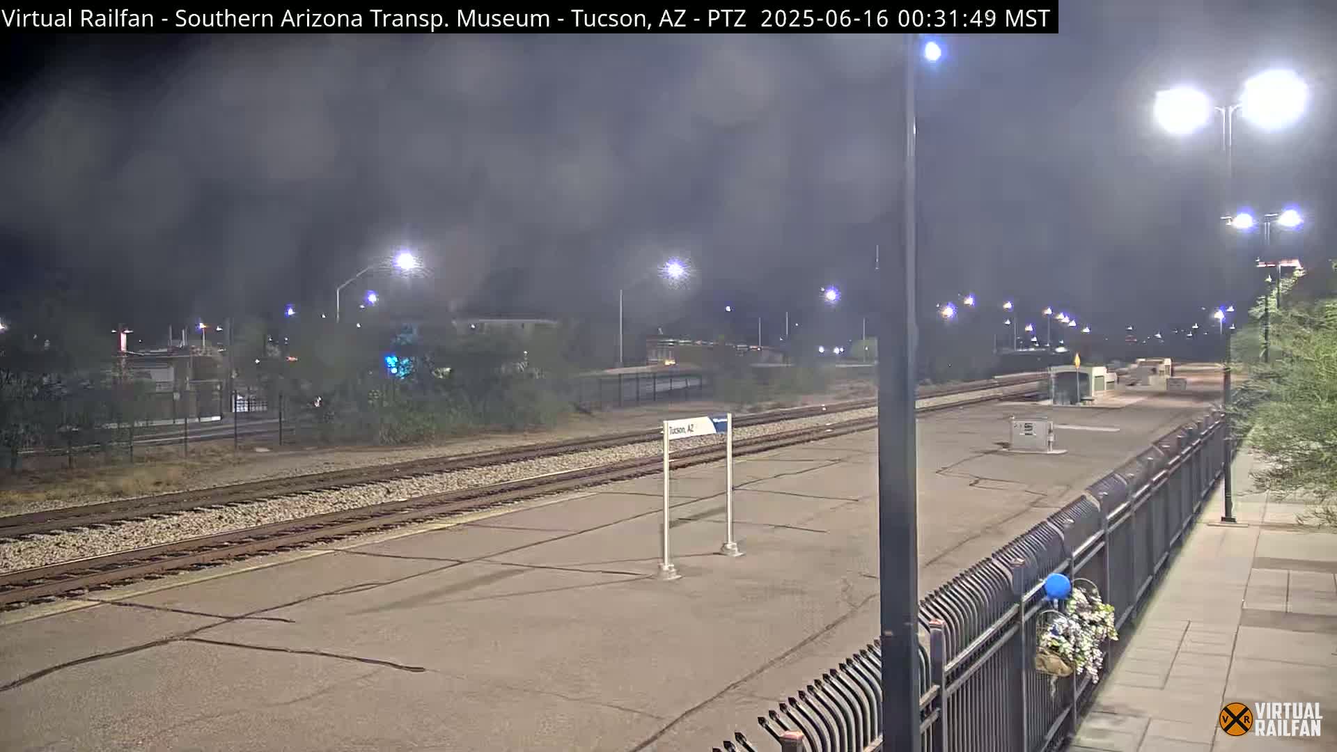 A nighttime view of a train station platform with train tracks, a fence, and streetlights under hazy conditions.