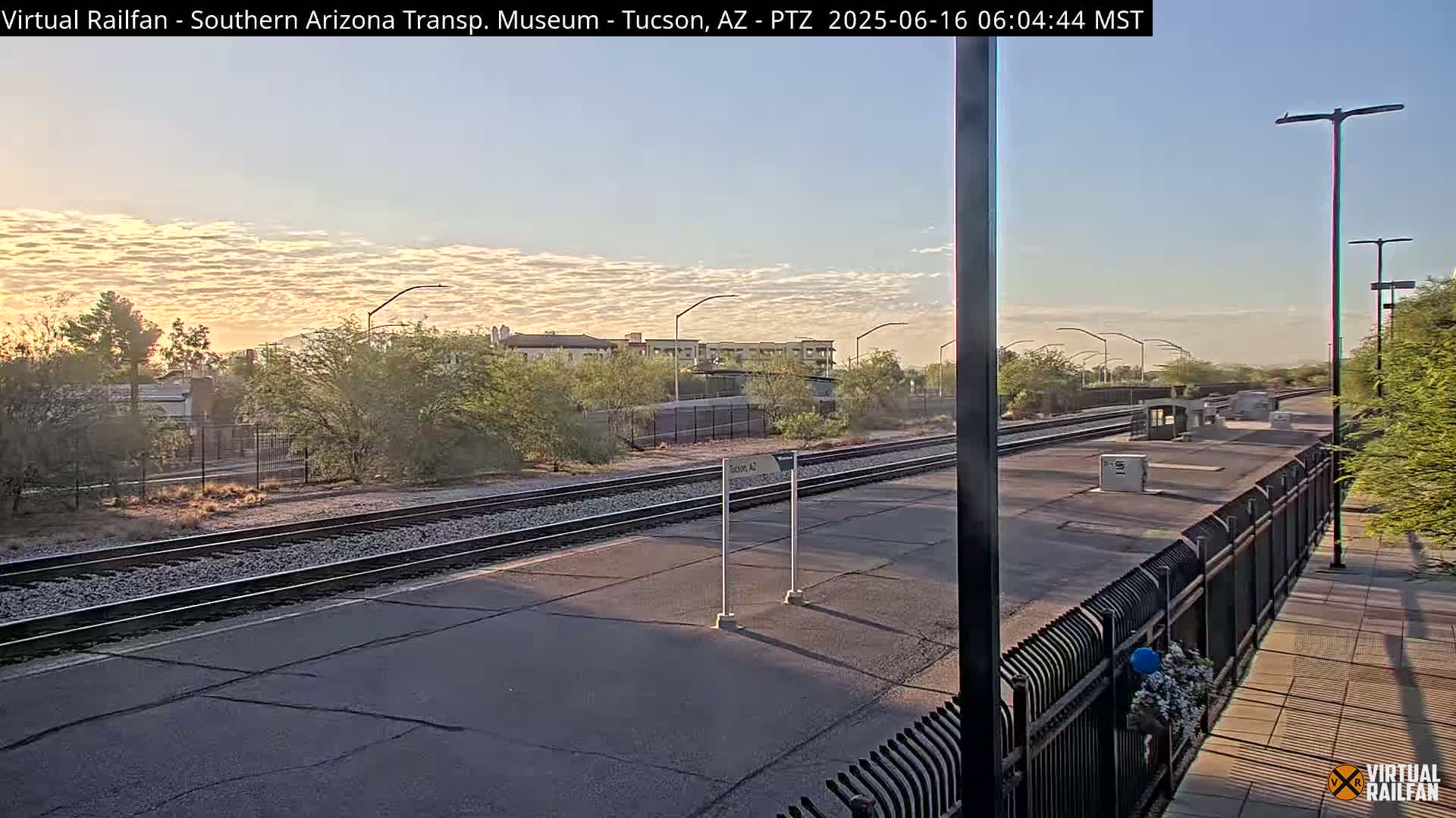 A sunrise illuminates a train station platform with a set of train tracks and a black metal fence separating the platform from a sidewalk.