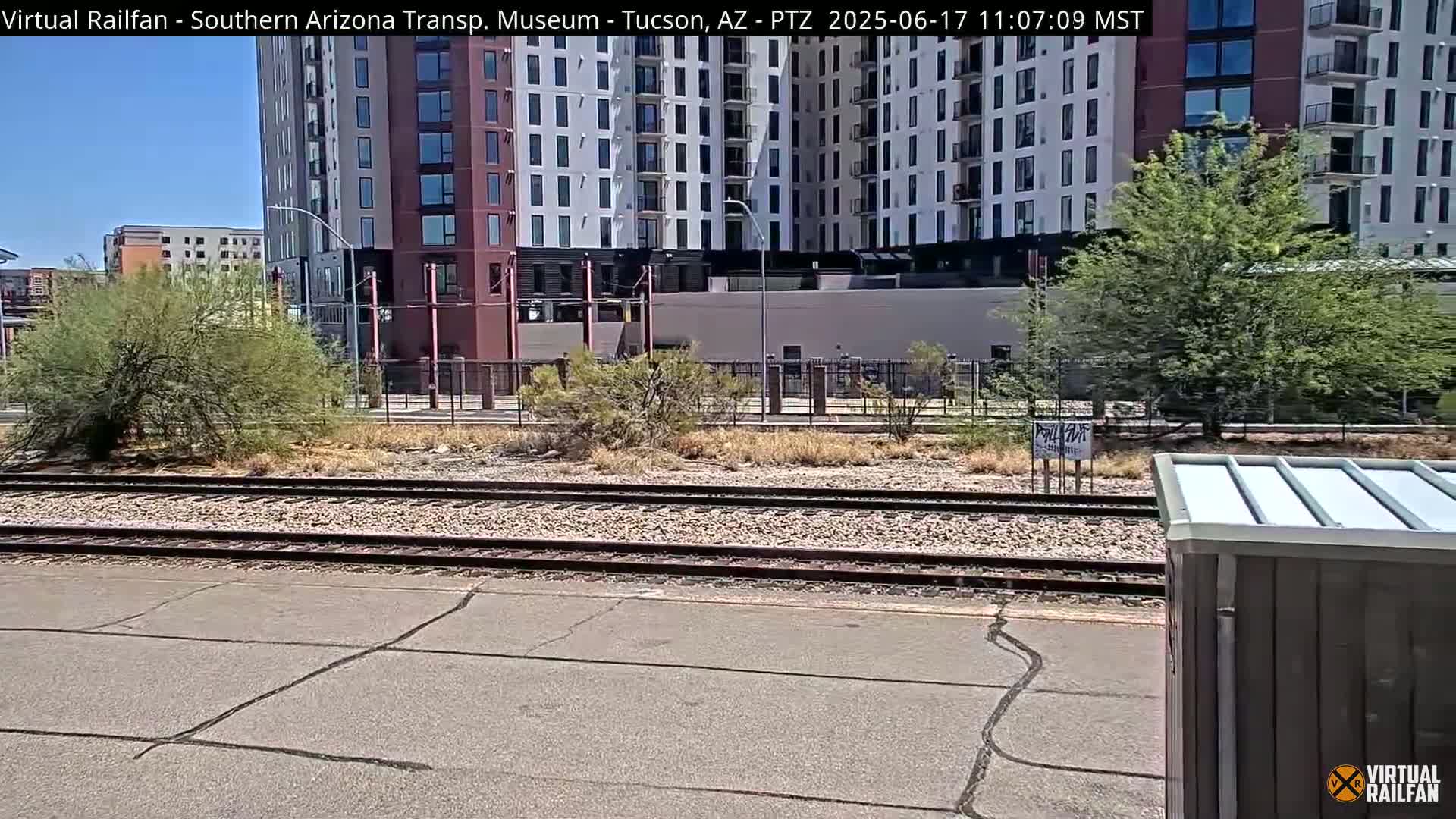 A sunny day view of railroad tracks in front of a tall apartment building and some desert vegetation.
