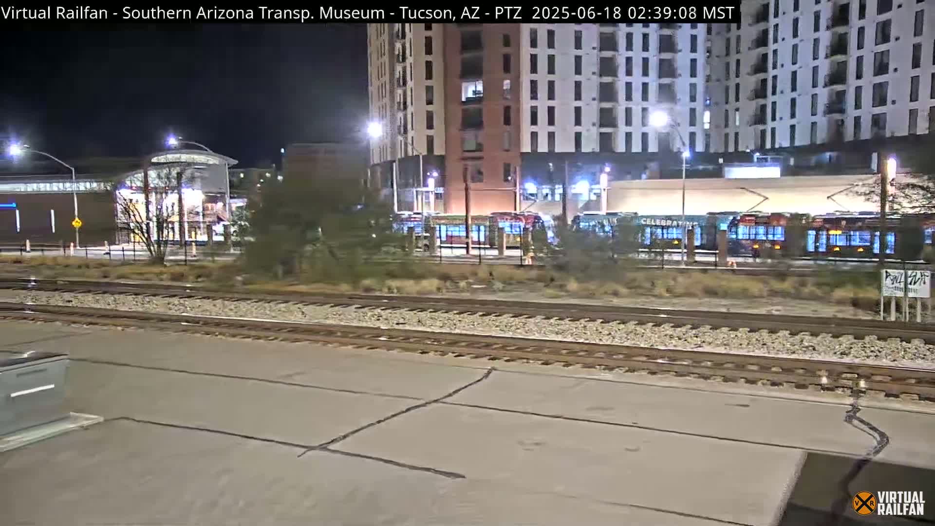 At night, several light-rail vehicles are parked near a modern building and a transit station next to train tracks.