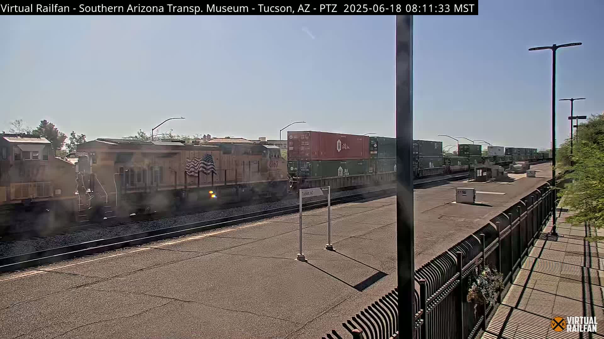A freight train, partially obscured by smoke, sits alongside a platform on a sunny day.