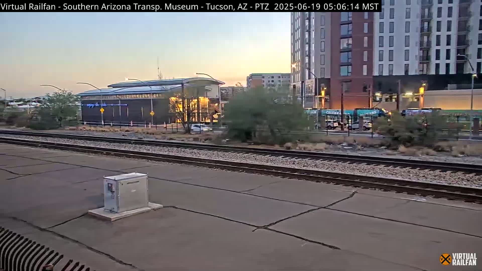Under a twilight sky, a train track runs beside a modern building and several light rail vehicles.