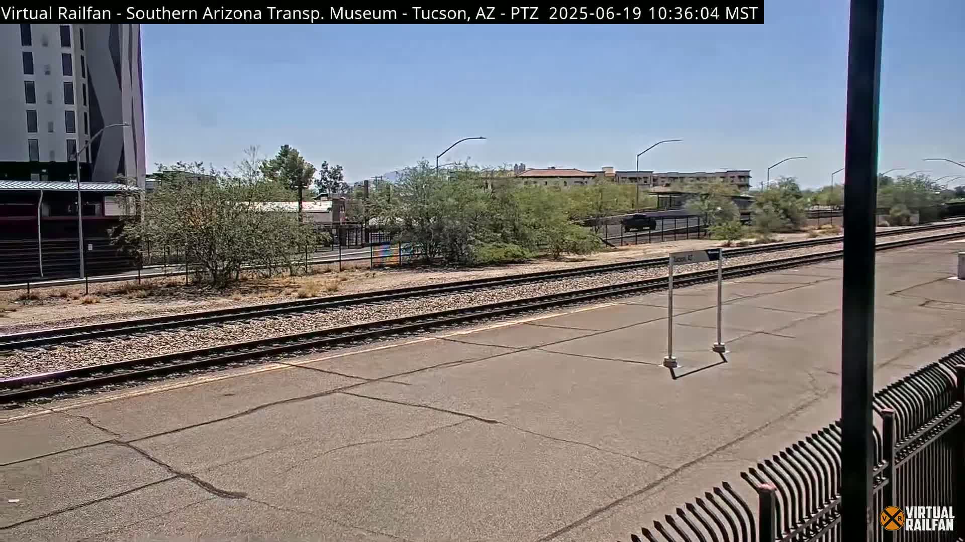 A sunny view of a train station platform with multiple train tracks, surrounded by desert landscaping and buildings in the distance.
