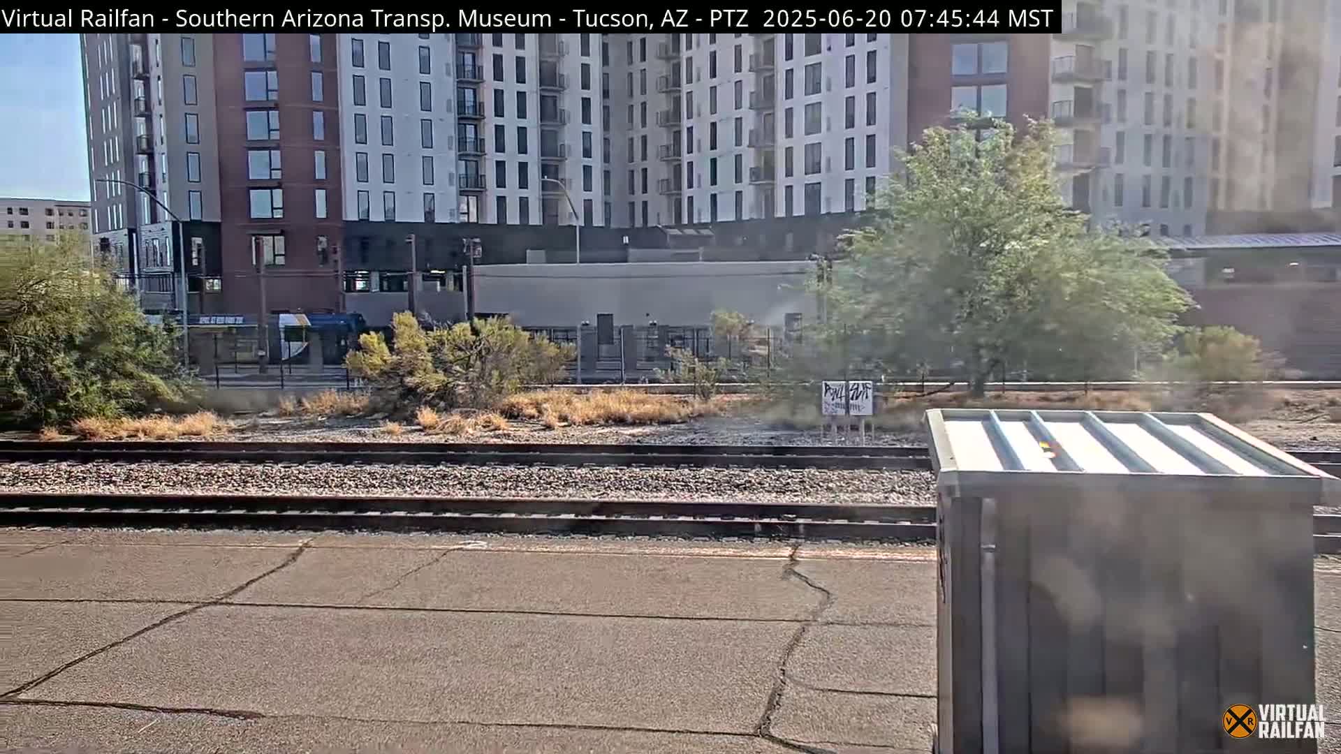 A view of train tracks next to a modern apartment building and sparse desert vegetation on a sunny day.