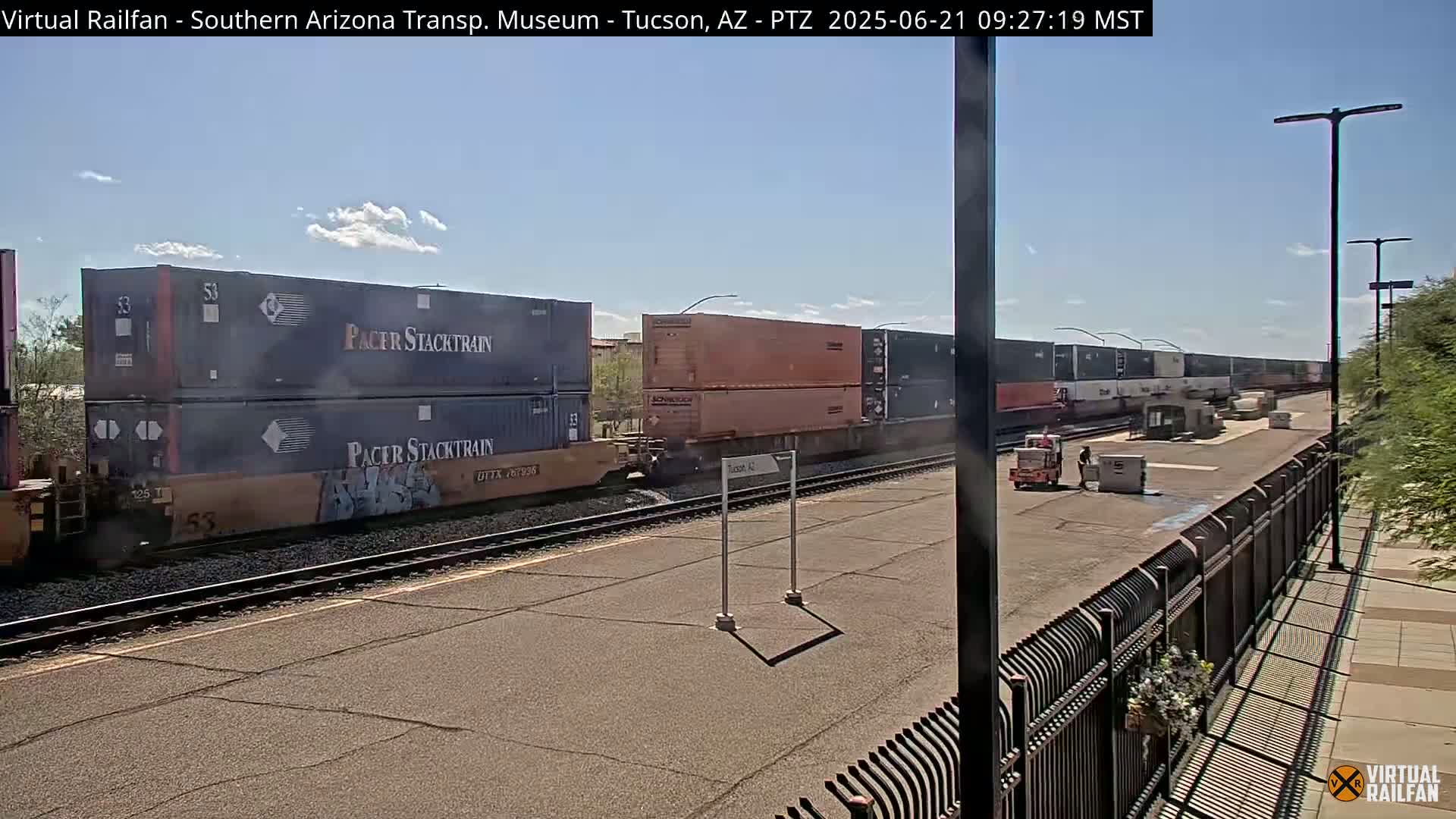 A long freight train carrying multiple shipping containers sits on railroad tracks under a mostly sunny sky.